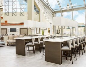 Bright atrium workspace with high-top communal tables, cushioned seating, floor-to-ceiling windows, and abundant natural light.
