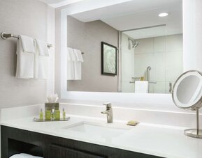 Contemporary bathroom vanity with large mirror, bright lighting, toiletries, towels, and glass-enclosed shower.
