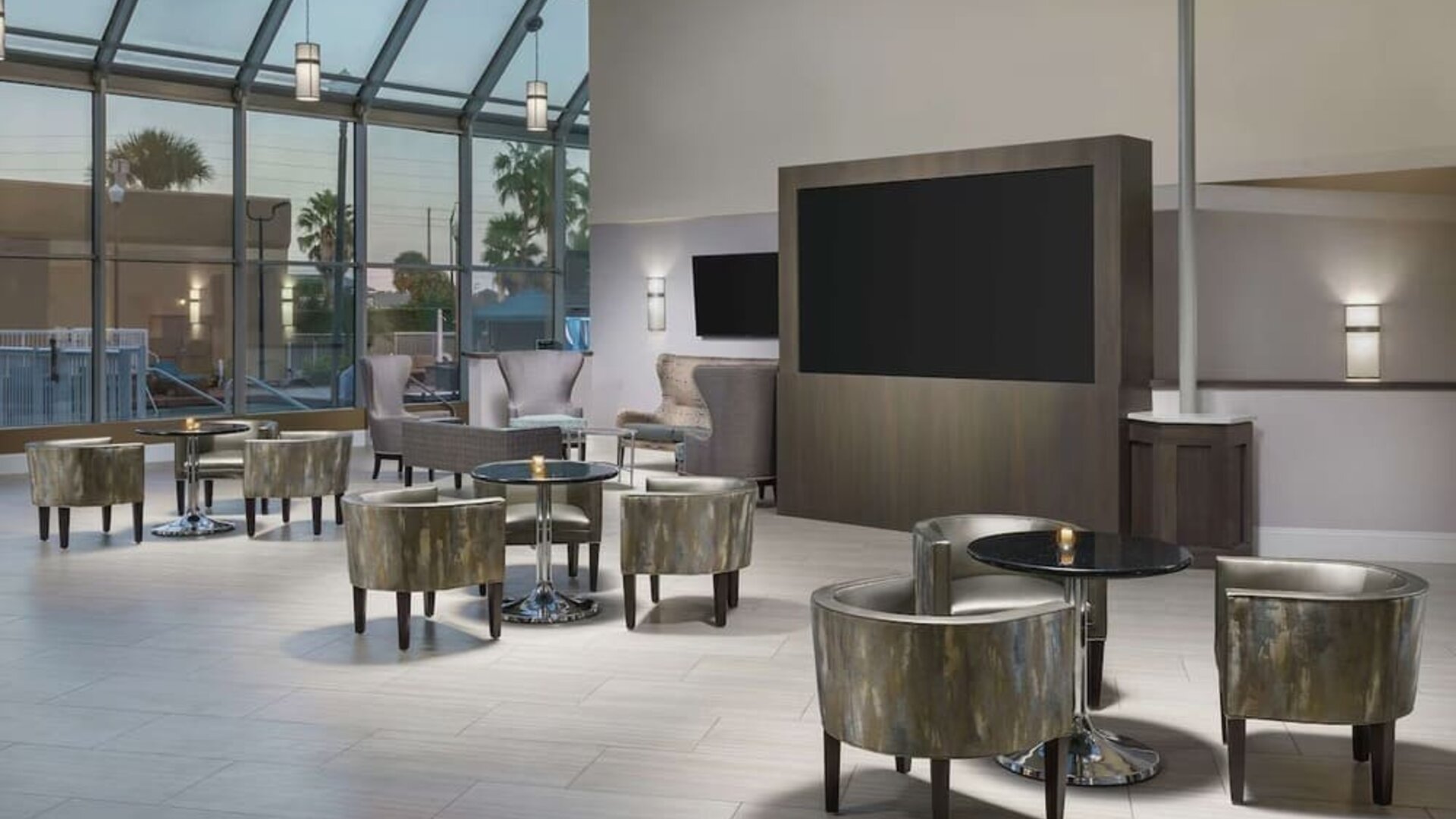 Elegant lobby lounge with armchairs, cocktail tables, soaring glass ceiling, and large display screen.