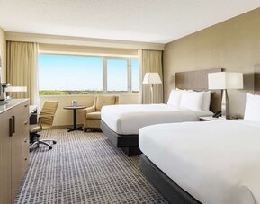 Guest room with two double beds, work desk, lounge chair, large window, and bright contemporary furnishings.