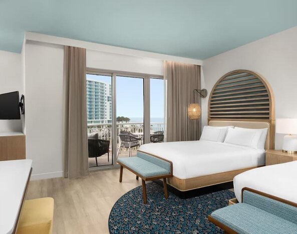 Guest room with two beds, patterned rug, balcony access, and views toward the beach and nearby buildings.