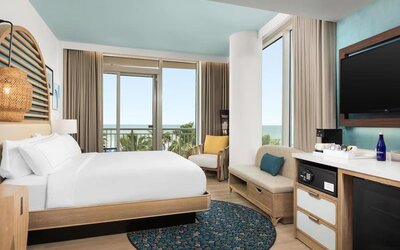 Contemporary guest room with king bed, seating area, large TV, and balcony offering partial ocean views.