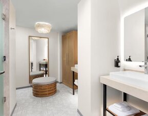 Spacious bathroom and dressing area with illuminated mirror, vessel sink, woven ottoman, and wood wardrobe.