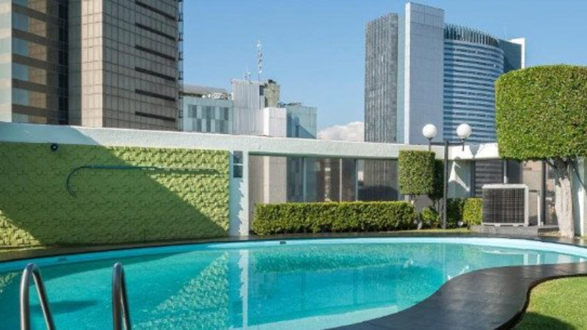 Outdoor swimming pool surrounded by landscaped greenery with high-rise city buildings beyond at Hotel Casa Blanca.