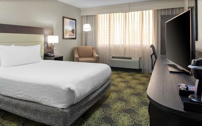 King guest room with plush bed, HDTV, armchair, workspace, and large windows providing natural daylight.
