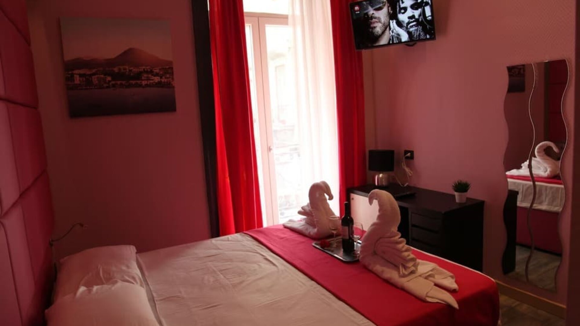 Double room with red curtains, balcony access, towel swans and wall-mounted TV at Dolce Vita Rooms & Breakfast.