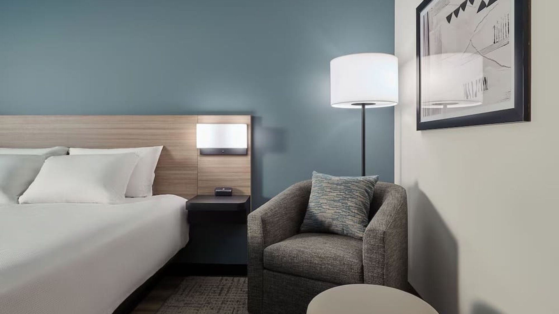 Cozy guest room corner with a plush armchair, ottoman, floor lamp and comfortable king bed at Hyatt Select Philadelphia Airport.