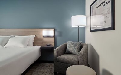 Cozy guest room corner with a plush armchair, ottoman, floor lamp and comfortable king bed at Hyatt Select Philadelphia Airport.