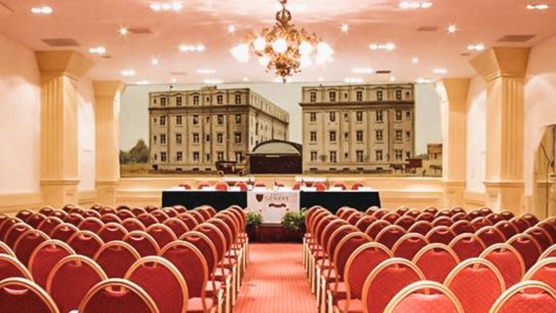 Large conference hall with rows of red chairs, a presentation stage, chandeliers, and a decorative mural backdrop at Hotel Geneve.
