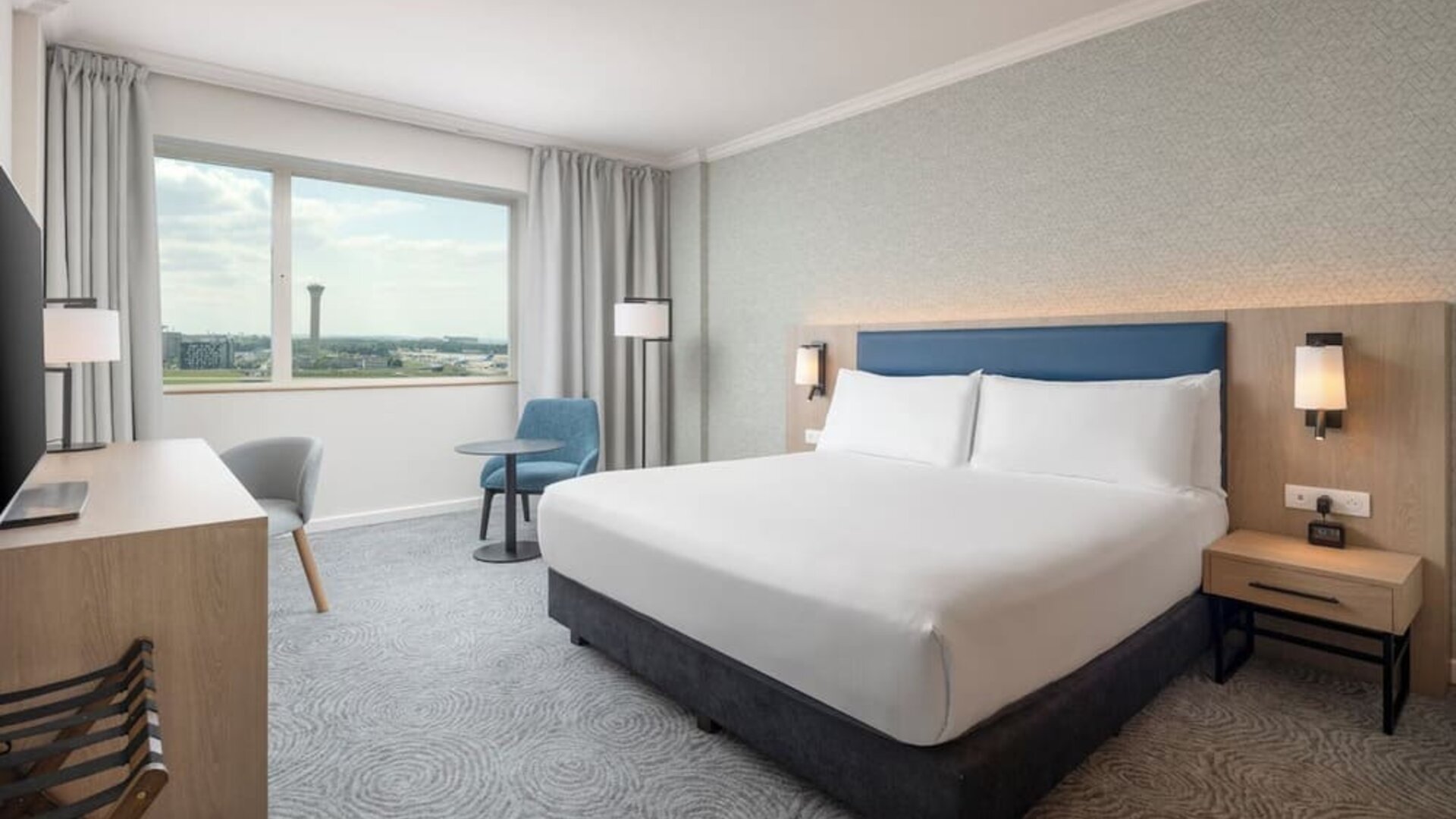 Modern king guest room with bed, large television, long work desk, armchairs, side table and floor-to-ceiling curtains.