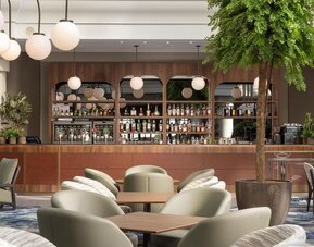 Contemporary hotel bar with upholstered lounge seating, wooden tables, globe pendant lights, plants and stocked bar.