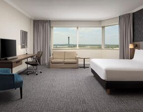 Spacious hotel room with king bed, sofa, work desk, television, armchair and panoramic windows overlooking the airport.