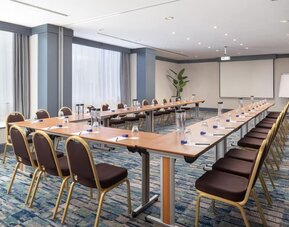 Hotel meeting room with U-shaped conference tables, chairs, presentation screen, flip chart and bottled water.