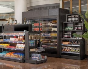 Hotel convenience store with refrigerated drinks, snacks, wine, toiletries and travel essentials displayed on shelves.