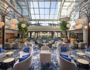 Expansive hotel lobby lounge beneath a glass atrium with sofas, armchairs, tables, blue carpet and globe lighting.