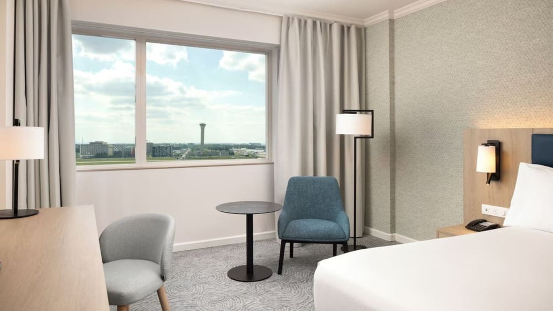 Spacious king guest room with large airport-view window, work desk, armchairs, side table and contemporary furnishings.