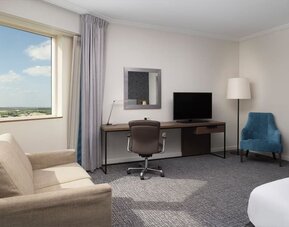 Spacious guest room workspace with desk, office chair, television, sofa, armchair and large window overlooking the airport. 