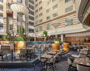 Large hotel atrium with restaurant tables, lounge seating, indoor trees, decorative columns and soaring glass ceiling