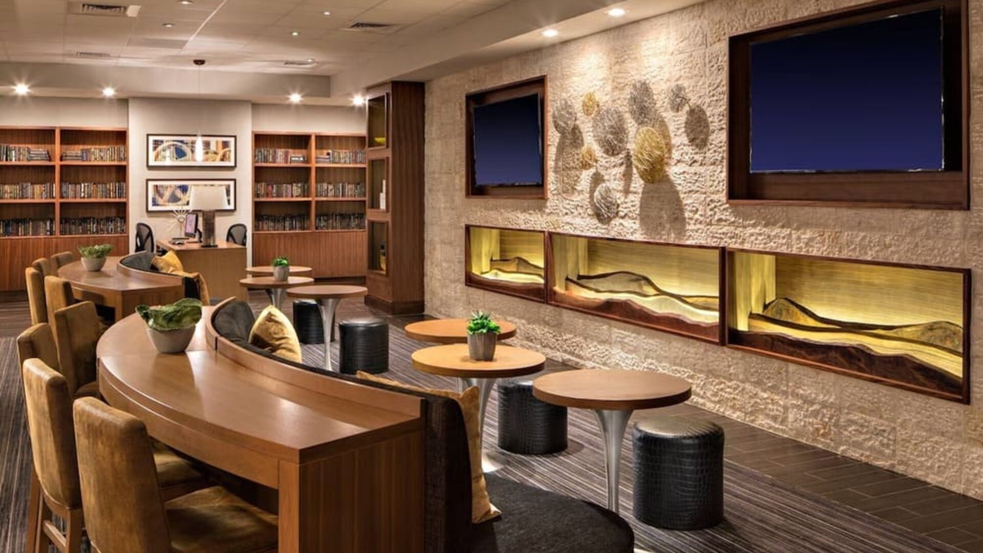 Lobby lounge featuring communal worktables, comfortable seating, built-in shelving, and a contemporary gathering space at Houston Marriott South at Hobby Airport.