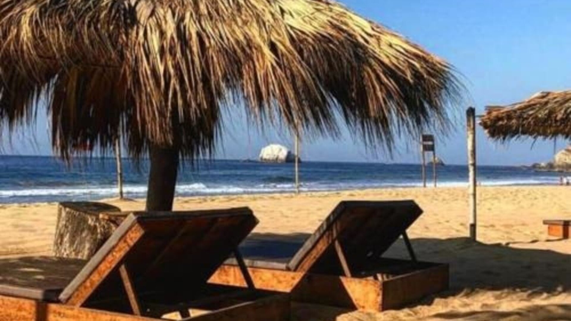 Two wooden sun loungers rest beneath a large thatched palapa on a sandy beach, overlooking the blue ocean and breaking waves under a clear sky.