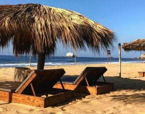 Two wooden sun loungers rest beneath a large thatched palapa on a sandy beach, overlooking the blue ocean and breaking waves under a clear sky.