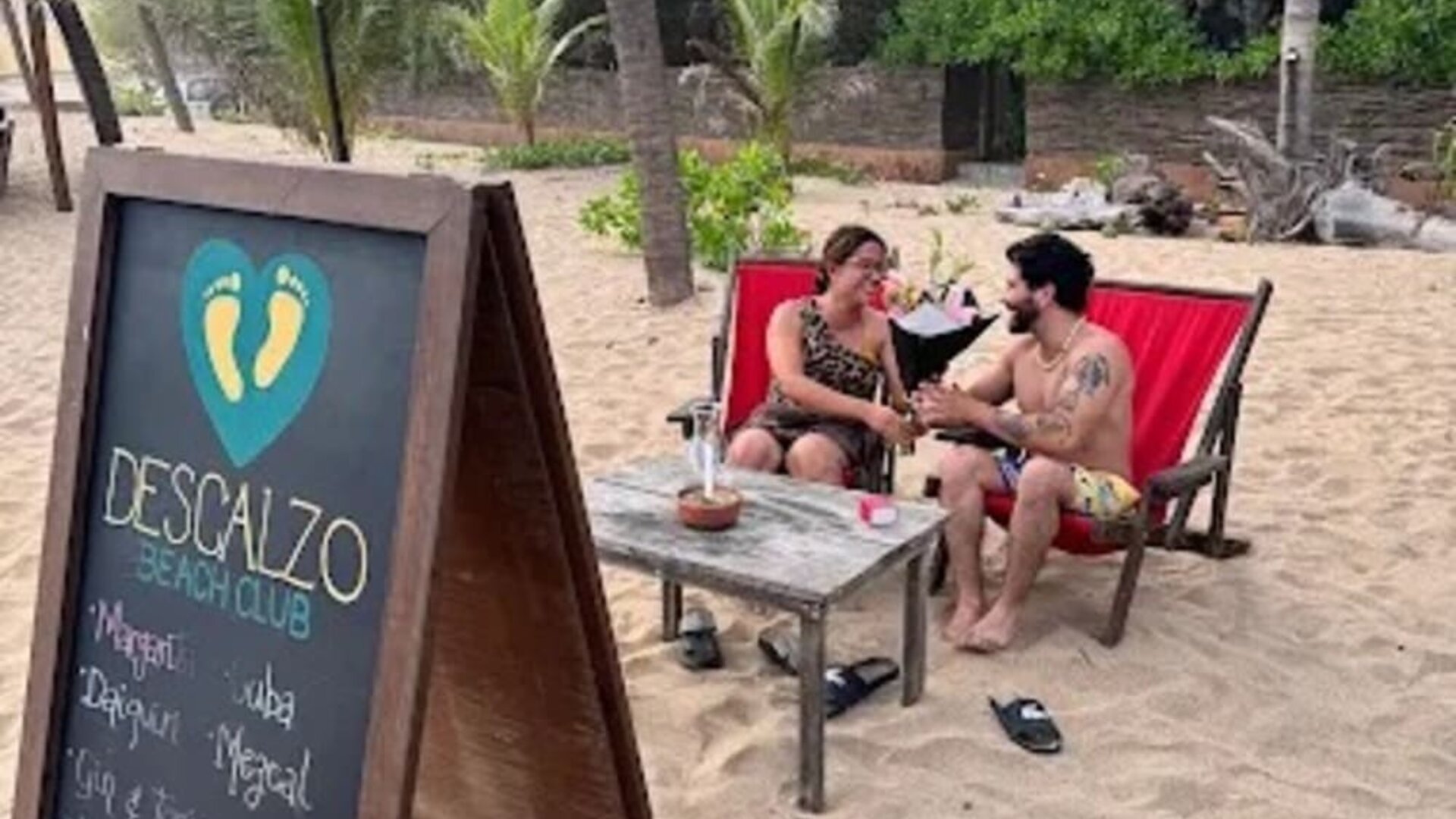 Descanso Beach Club sign beside two guests relaxing in red lounge chairs on the sand, surrounded by palm trees, tropical greenery, and casual beachfront seating.