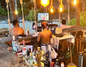 Rustic wooden beach bar with a large selection of liquor bottles, warm pendant lights, and staff behind the counter, with palm trees and an outdoor television beyond.