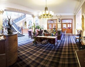 Grand hotel lobby featuring tartan carpeting, elegant seating, a sweeping staircase, and traditional Scottish décor at The Royal Highland Hotel.