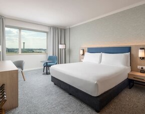 Modern king guest room with bed, large television, long work desk, armchairs, side table and floor-to-ceiling curtains.