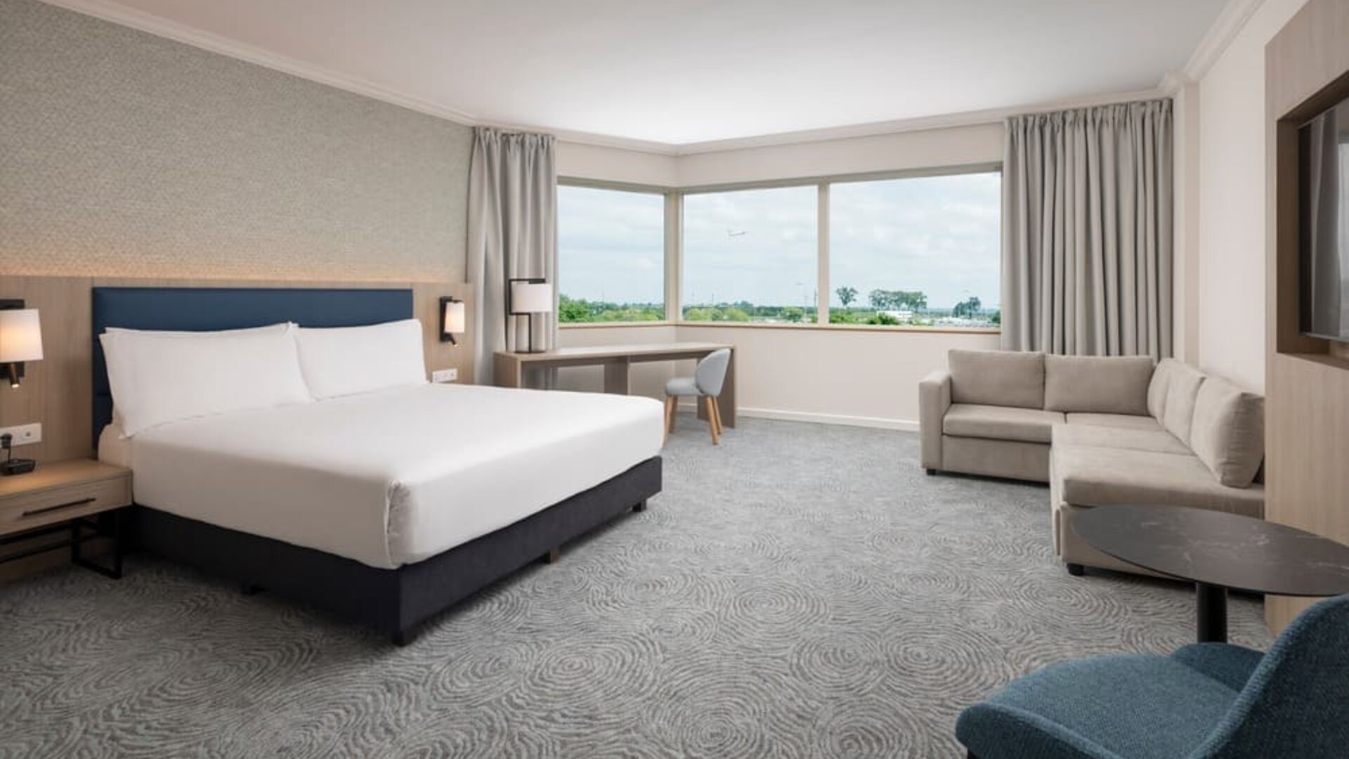 Spacious king guest room with large airport-view window, work desk, armchairs, side table and contemporary furnishings.