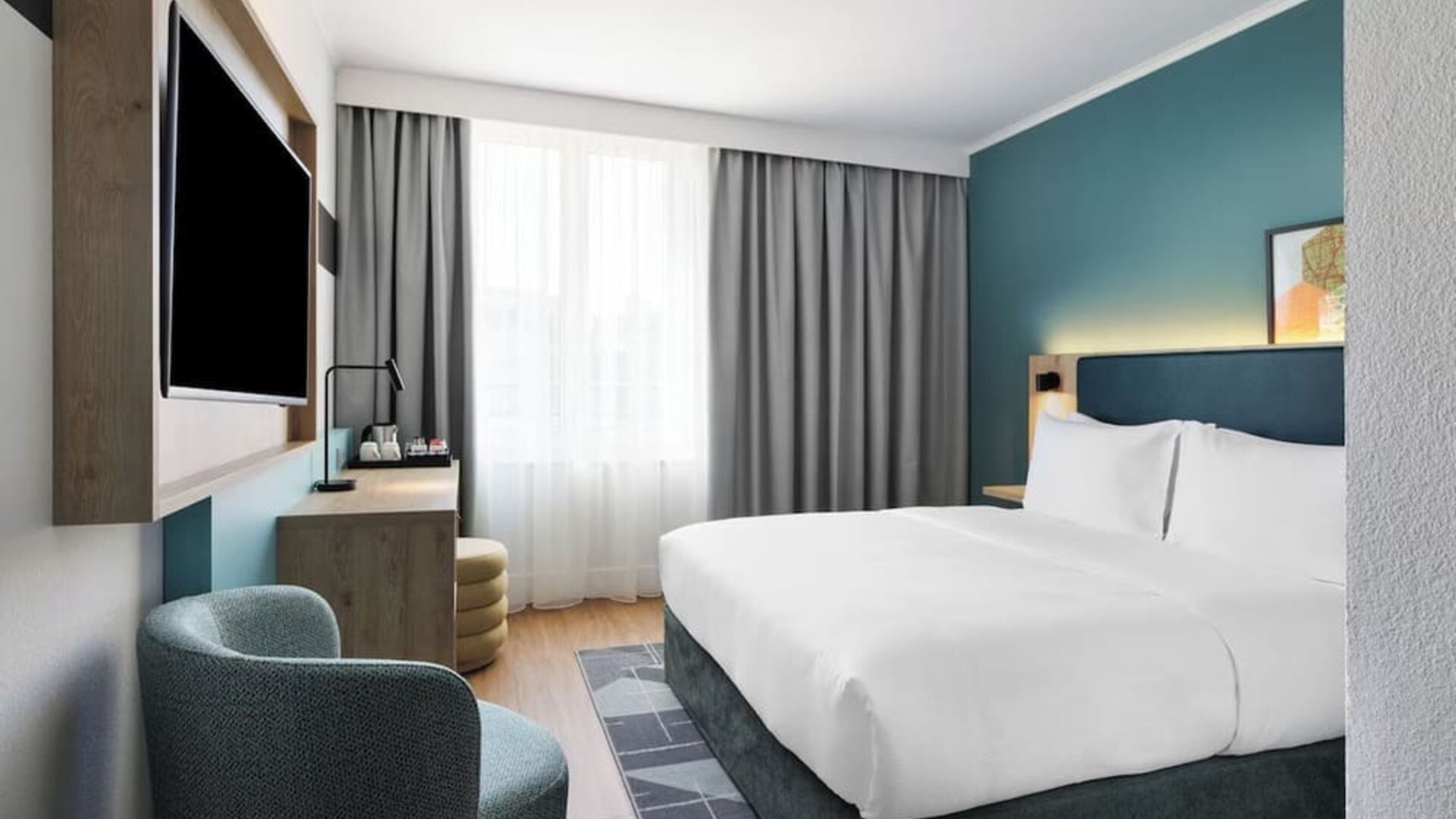 Bright contemporary hotel room with a king bed, white bedding, teal accent wall, large window with gray curtains, wall-mounted TV, desk, and upholstered armchair.