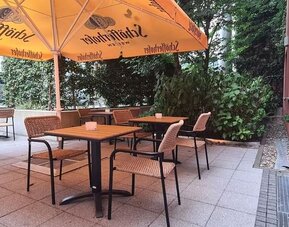 Shaded hotel terrace with wooden tables, woven chairs, large orange umbrellas, tiled flooring, and dense green trees and shrubs surrounding the seating area.