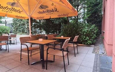 Shaded hotel terrace with wooden tables, woven chairs, large orange umbrellas, tiled flooring, and dense green trees and shrubs surrounding the seating area.