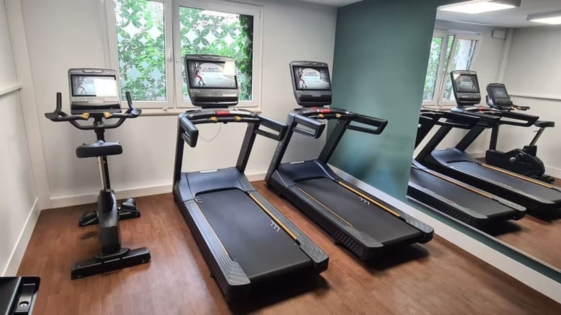Compact hotel fitness center with two treadmills, a stationary exercise bike, mirrored wall, wood-look flooring, and windows providing natural light.