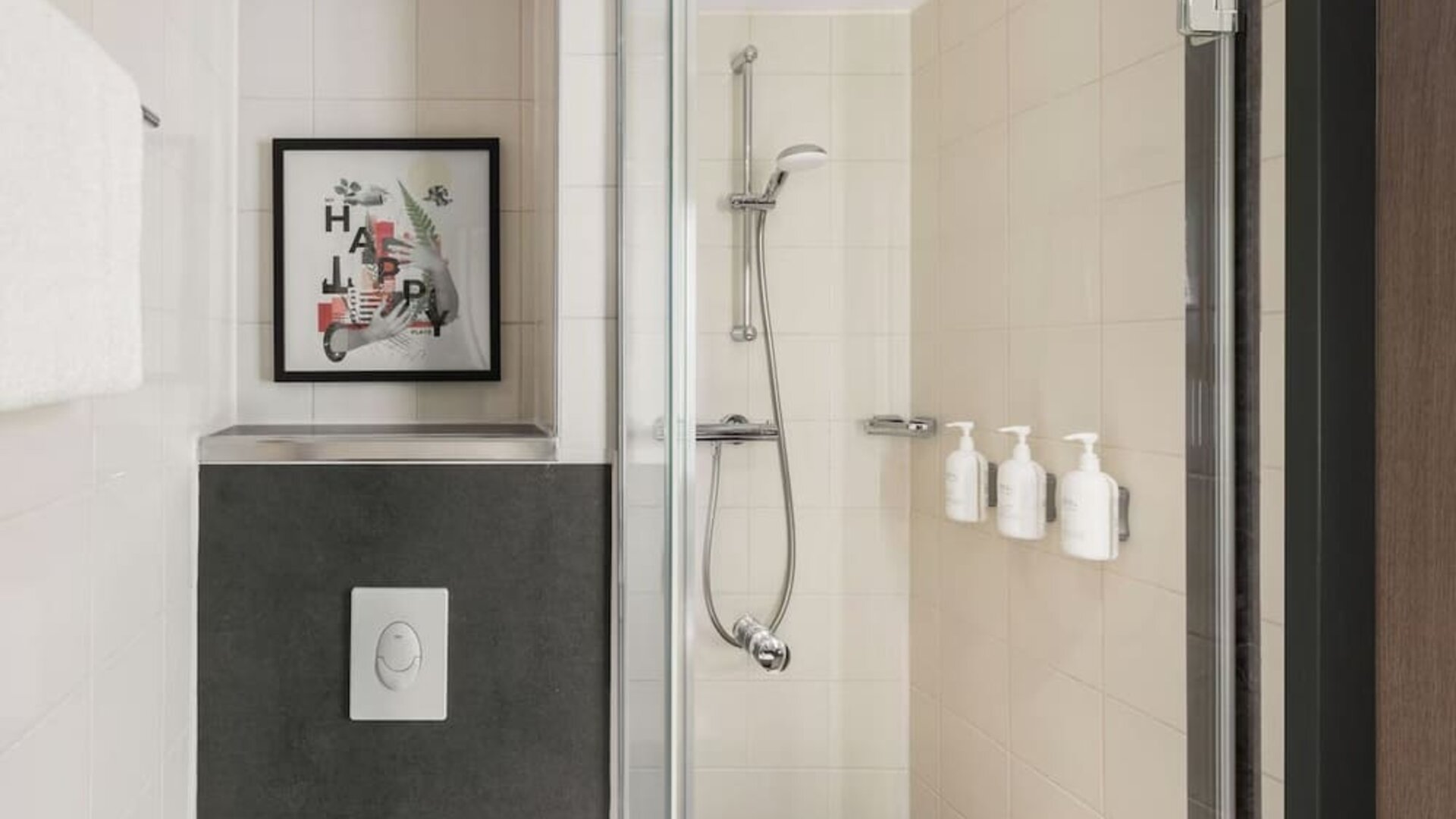 Modern tiled hotel bathroom featuring a glass-enclosed walk-in shower with handheld showerhead, wall-mounted toiletries, white towels, and framed artwork.