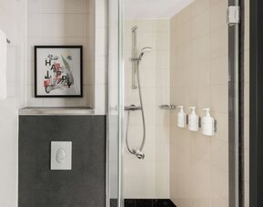 Modern tiled hotel bathroom featuring a glass-enclosed walk-in shower with handheld showerhead, wall-mounted toiletries, white towels, and framed artwork.