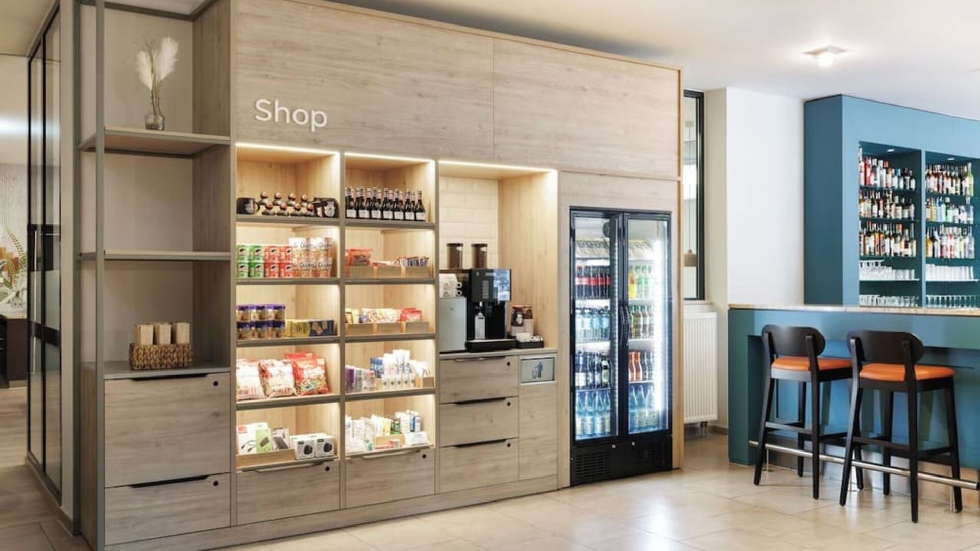 Hotel lobby shop with illuminated wooden shelves displaying snacks and drinks, a coffee machine, refrigerated beverages, and adjacent bar seating.