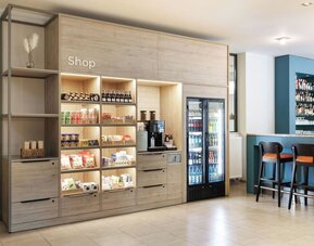 Hotel lobby shop with illuminated wooden shelves displaying snacks and drinks, a coffee machine, refrigerated beverages, and adjacent bar seating.