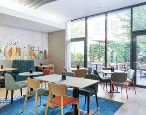 Contemporary hotel dining area with colorful upholstered chairs, wooden tables, teal booth seating, large floor-to-ceiling windows, and views of a leafy outdoor terrace.