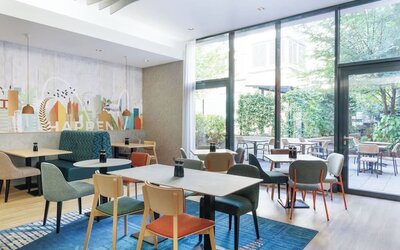 Contemporary hotel dining area with colorful upholstered chairs, wooden tables, teal booth seating, large floor-to-ceiling windows, and views of a leafy outdoor terrace.