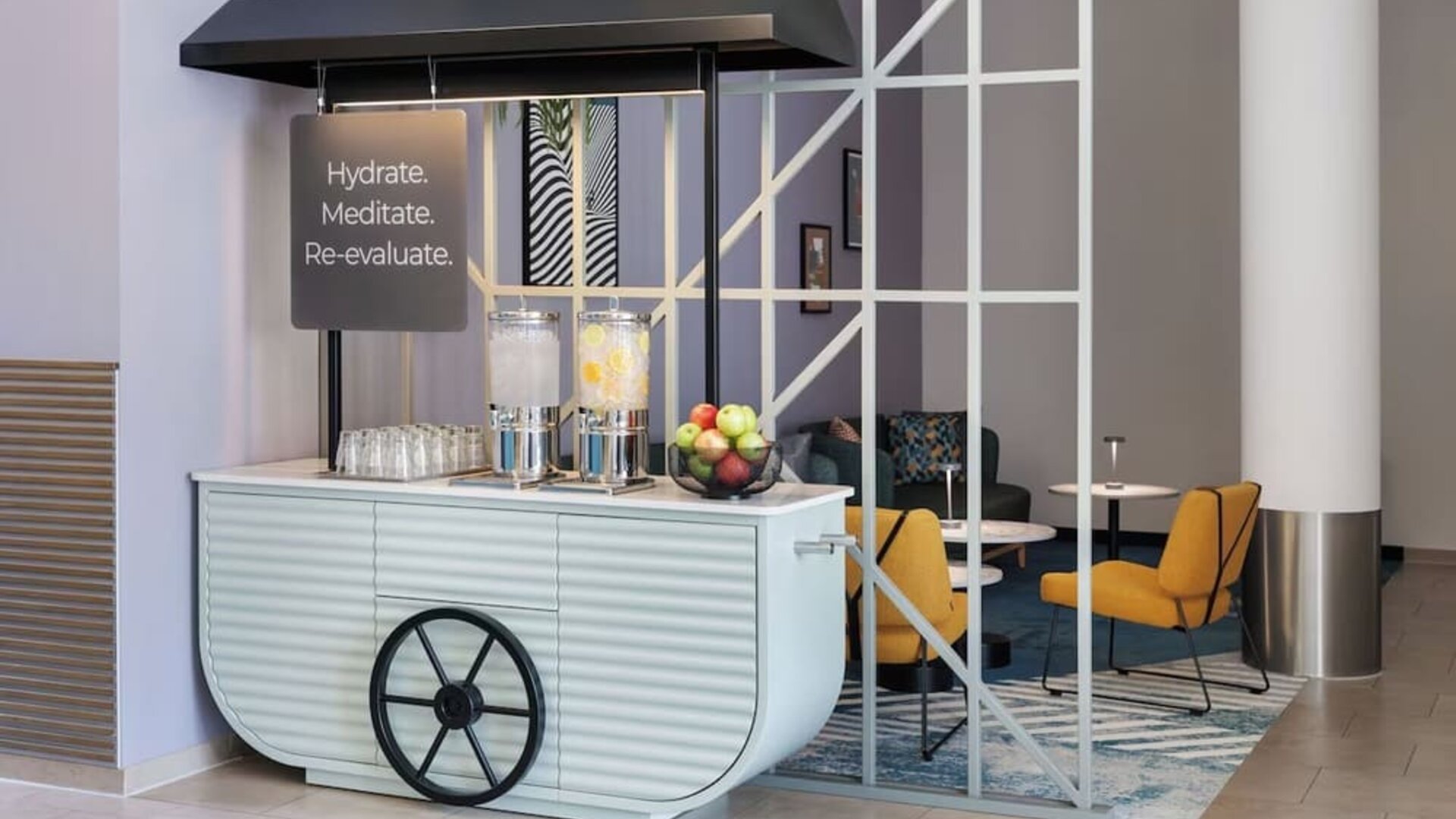 Stylish lobby hydration station designed like a cart, with two glass water dispensers, drinking glasses, a bowl of fresh fruit, and nearby lounge seating.