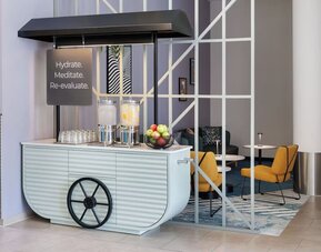 Stylish lobby hydration station designed like a cart, with two glass water dispensers, drinking glasses, a bowl of fresh fruit, and nearby lounge seating.