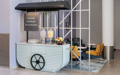Stylish lobby hydration station designed like a cart, with two glass water dispensers, drinking glasses, a bowl of fresh fruit, and nearby lounge seating.