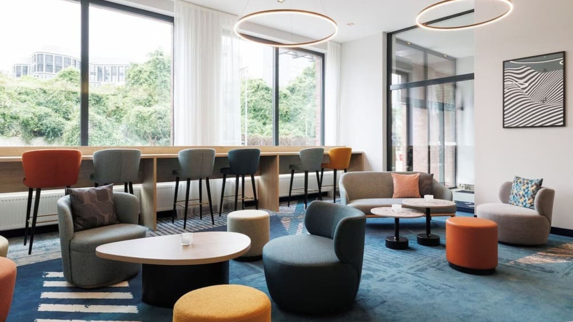 Spacious hotel lounge with teal, gray, orange, and yellow seating arranged around low tables, a blue patterned rug, large windows, and circular pendant lights.