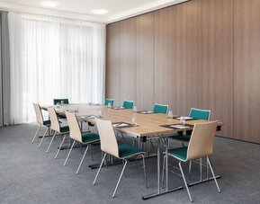 Bright hotel meeting room with rectangular tables arranged in a boardroom layout, teal and light wood chairs, water glasses and notepads, gray carpeting, and floor-to-ceiling curtains.