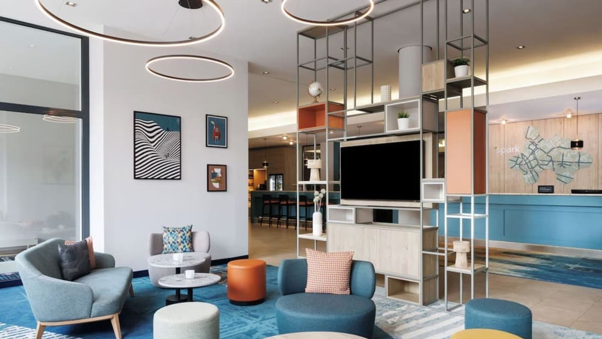 Contemporary hotel lobby with colorful lounge chairs and ottomans, round tables, blue patterned carpeting, circular ceiling lights, open shelving, and reception desk in the background.