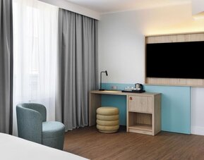 Hotel guest room workspace with wall-mounted TV, light wood desk, black reading lamp, electric kettle with cups, round upholstered stool, armchair, and floor-to-ceiling curtains.