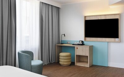 Hotel guest room workspace with wall-mounted TV, light wood desk, black reading lamp, electric kettle with cups, round upholstered stool, armchair, and floor-to-ceiling curtains.