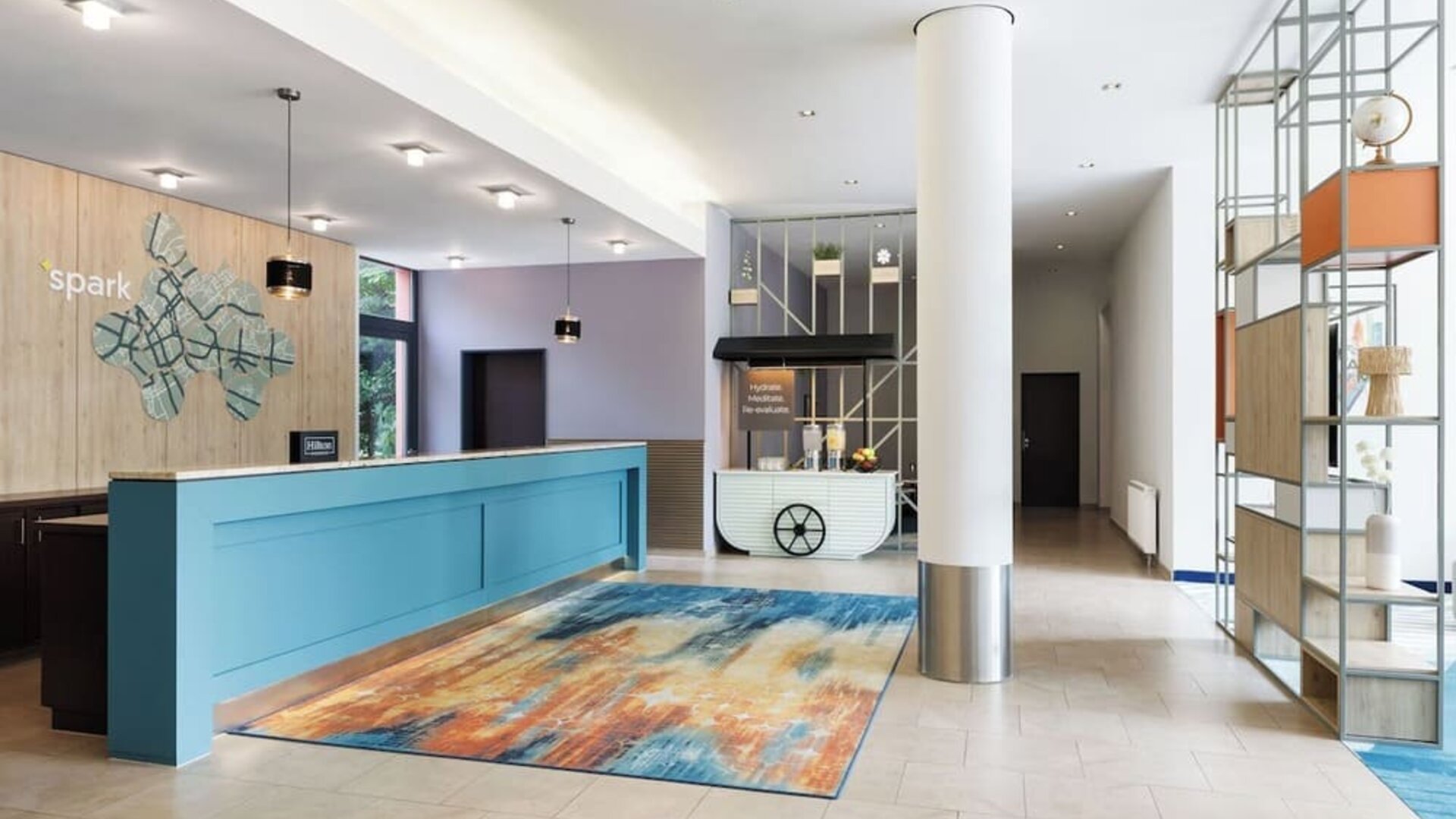 Modern hotel reception area with a turquoise front desk, colorful abstract rug, decorative open shelving, white column, hydration station, and Spark branding on a wood-paneled wall.