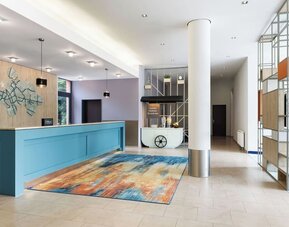 Modern hotel reception area with a turquoise front desk, colorful abstract rug, decorative open shelving, white column, hydration station, and Spark branding on a wood-paneled wall.